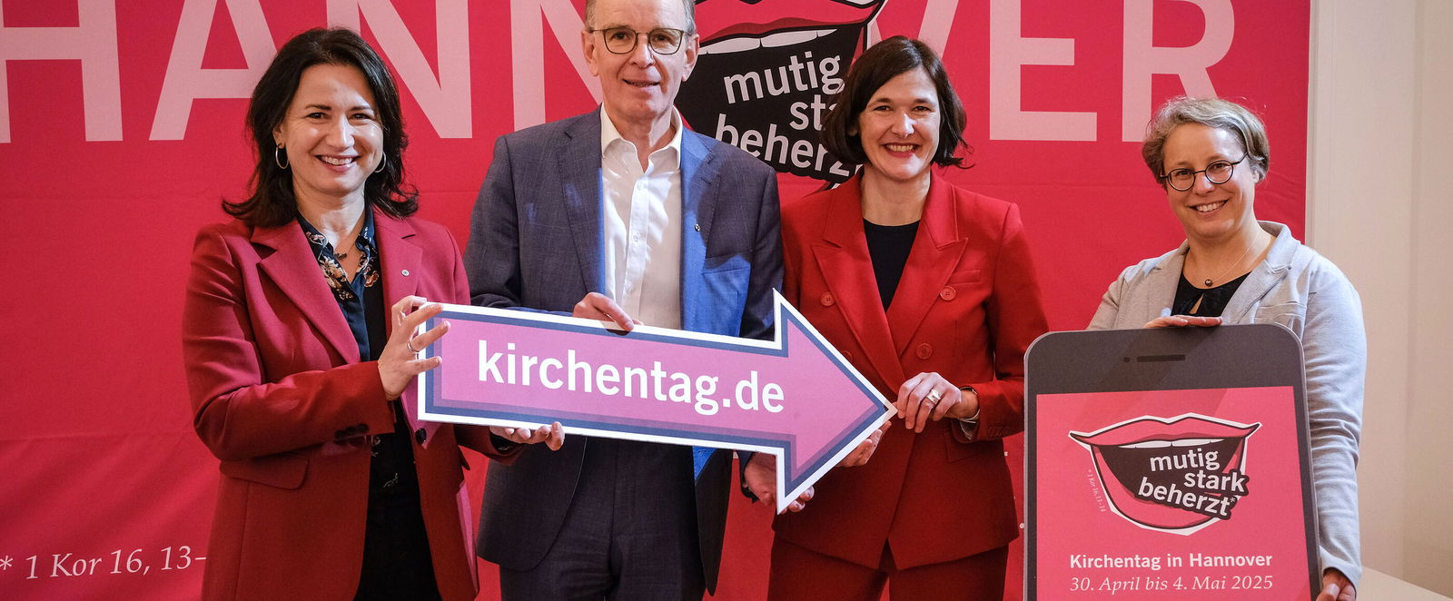 Drei als Frauen lesbare Personen und eine als Mann lesbare Person halten einen Pfeil mit der Aufschrift kirchentag.de und zeigen auf ein übergroßes Smartphone-Modell.