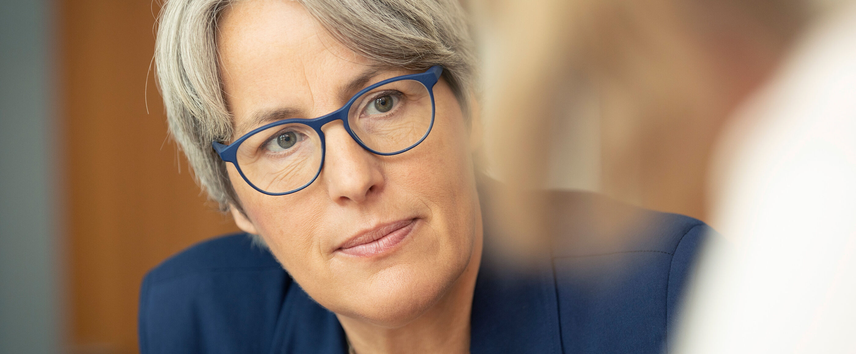 Eine als Frau lesbare Person mit blauer Brille und blauem Jacket hört einer anderen Person zu.