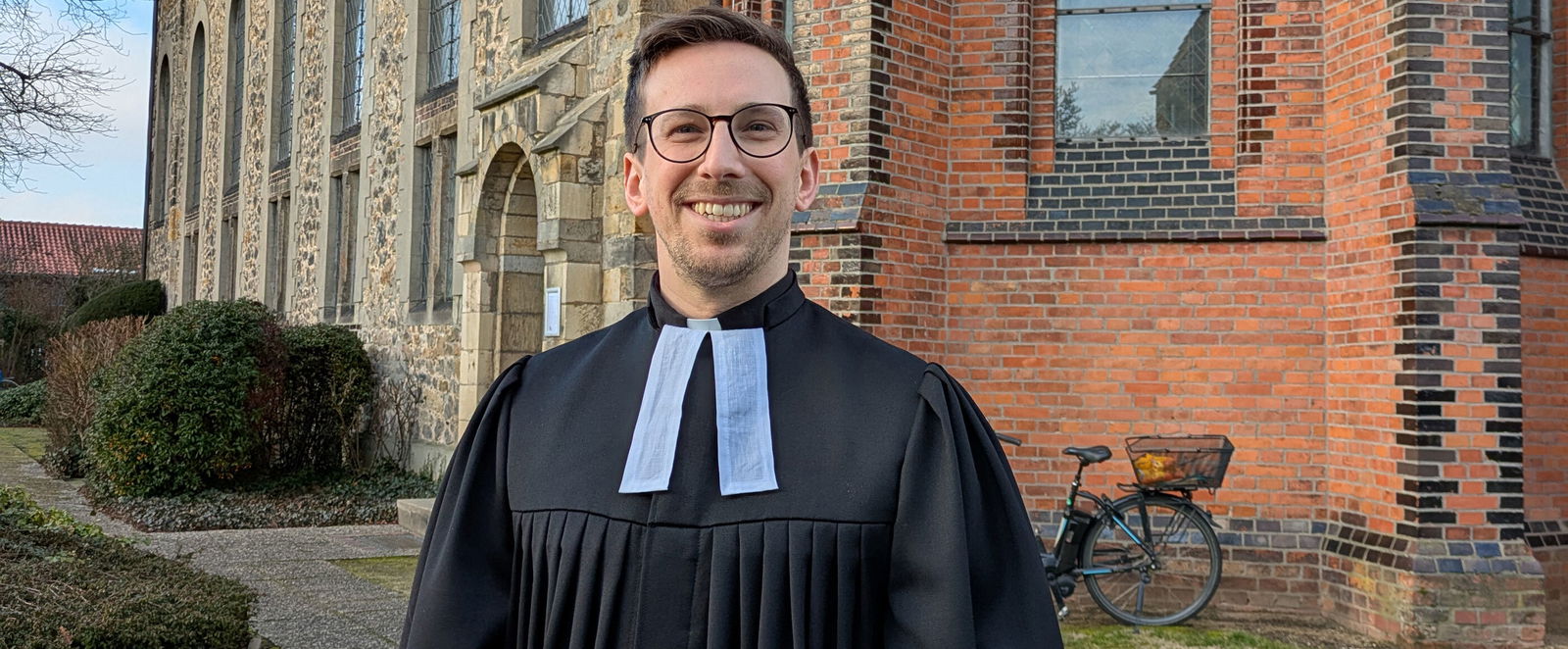 Eine als Mann lesbare Person im Talar steht vor einer Backsteinkirche.