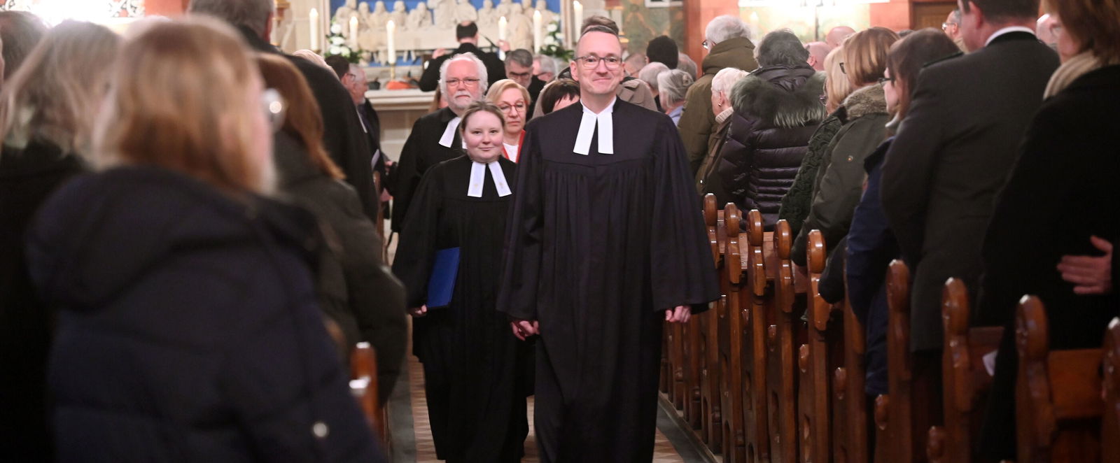 Eine männlich und eine weiblich gelesene Person, jeweils im Talar, gehen den Mittelgang einer vollbesetzten Kirche hinunter.