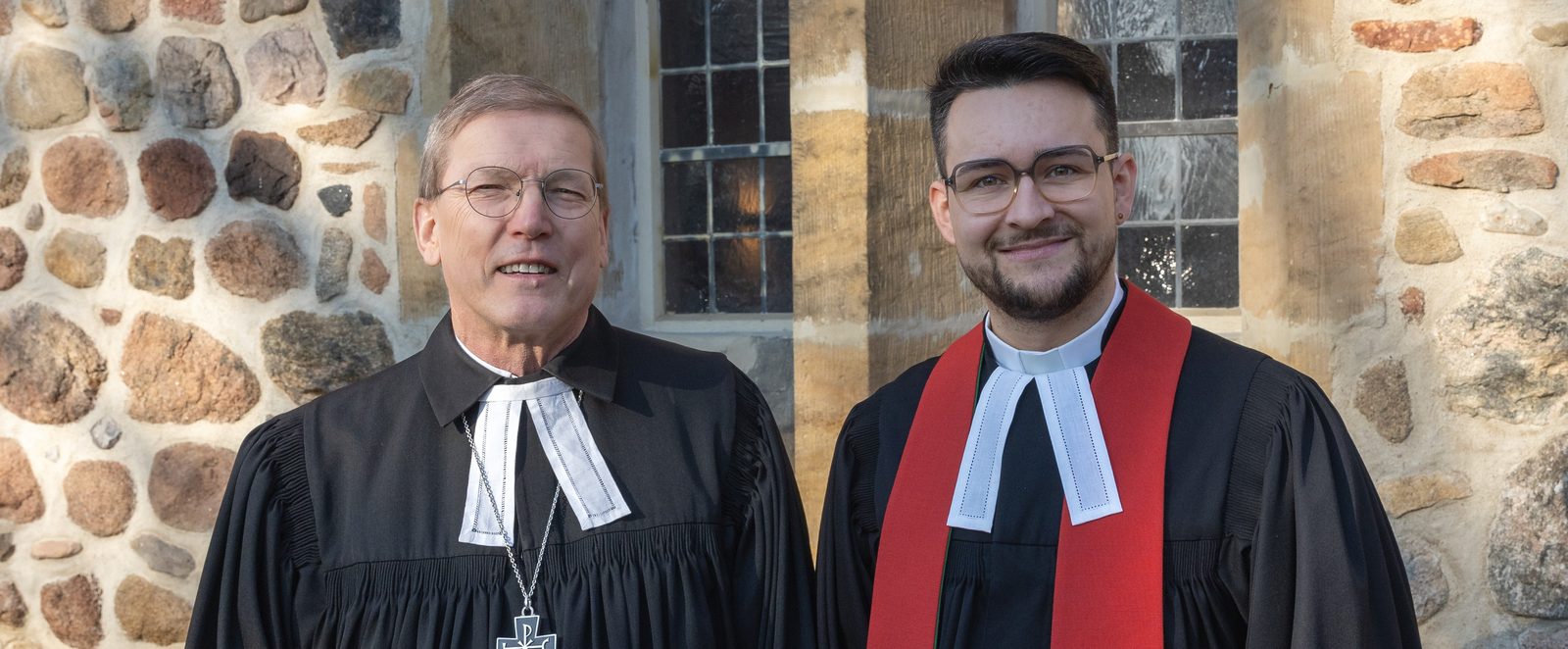 Zwei männlich gelesene Personen in Talaren stehen nebeneinander vor einer Steinwand, einer Kirche.
