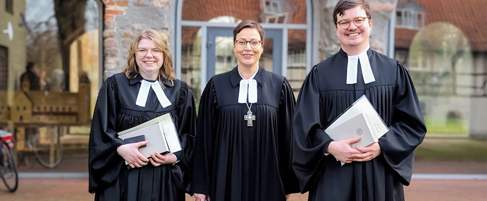 Zwei als Frauen lesbare Person und eine als Mann lesbare Person im Talar lächeln in die Kamera.