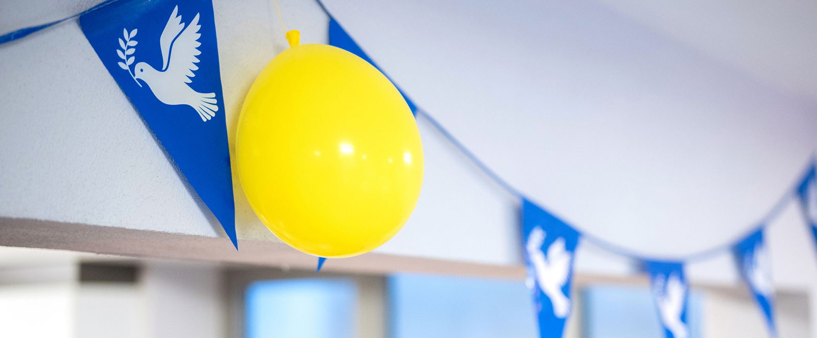 Eine blaue Wimpelkette mit Friedenstauben, dazwischen hängt ein gelber Luftballon.