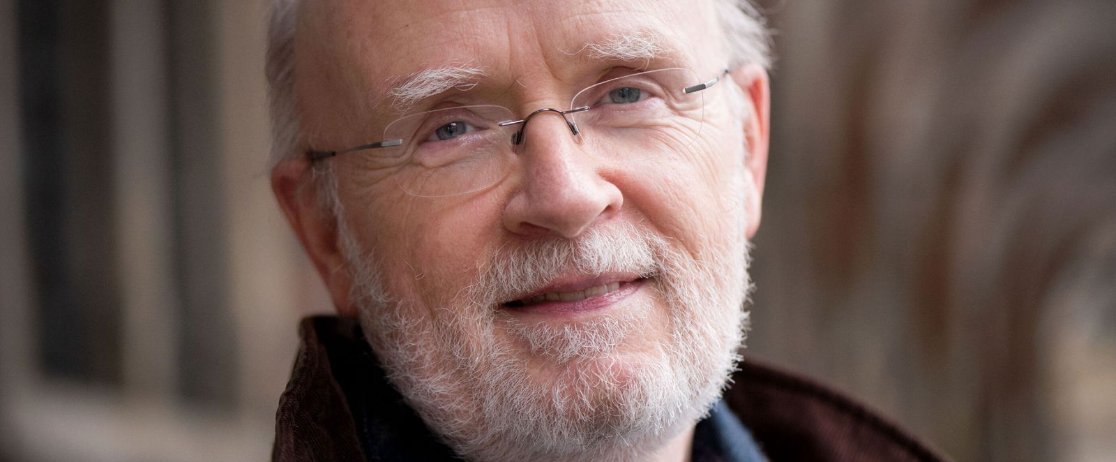 Eine als Mann lesbare Person mit hoher Stirn, grauem Bart und Brille