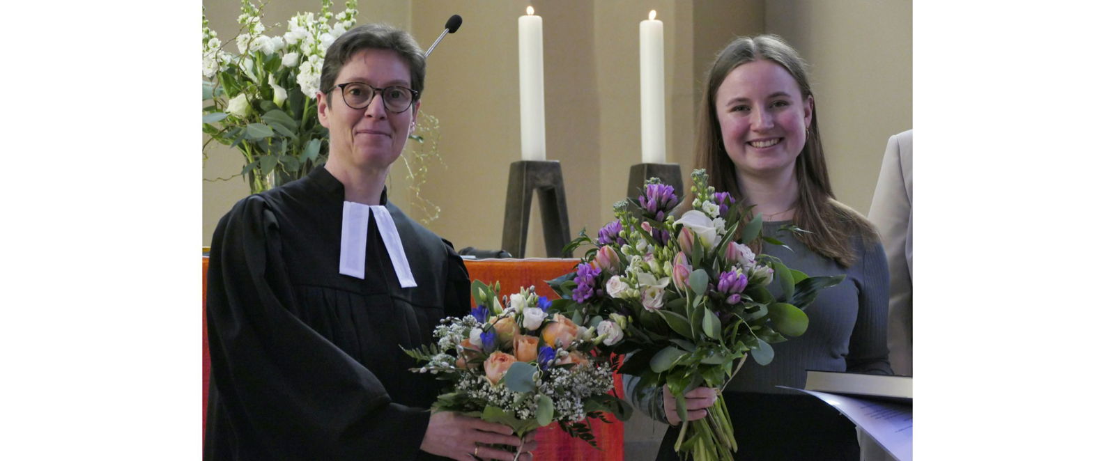 Zwei Frauen, eine mit Talar und eine andere mit Blumenstrauß.