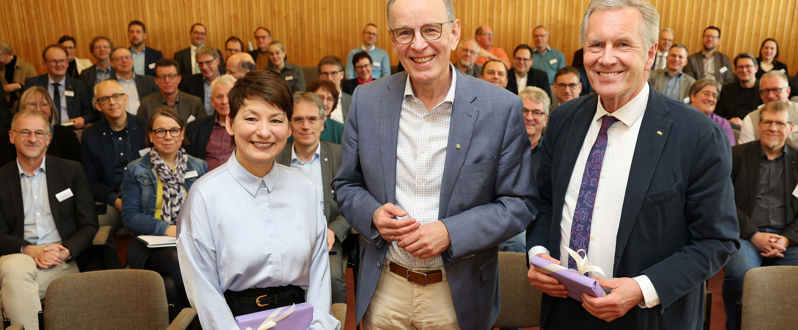 Eine als Frau lesbare Person und zwei als Männer lesbare Personen stehen vor einer größeren Gruppe in einem Tagungssaal.