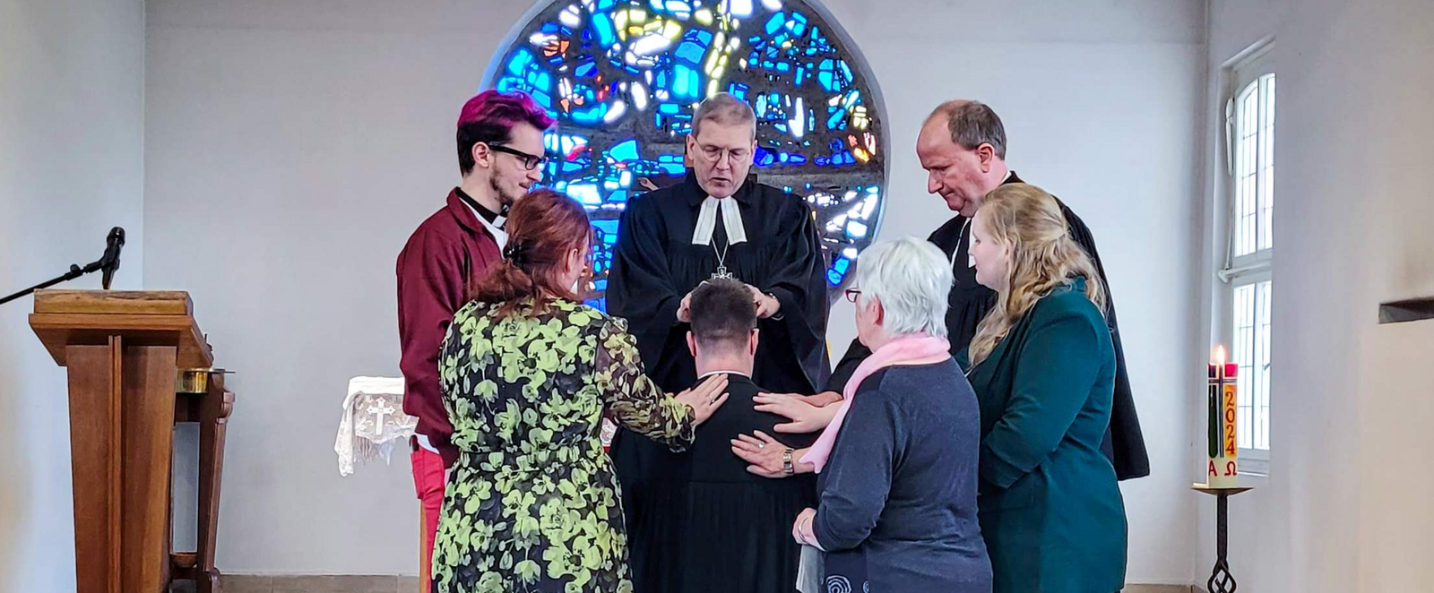Sechs Personen, einer in Talar, stehen ringsum eine knieende Person. Sie befinden sich in einem Altarraum mit einem blauen Buntglasfenster im Hintergrund.