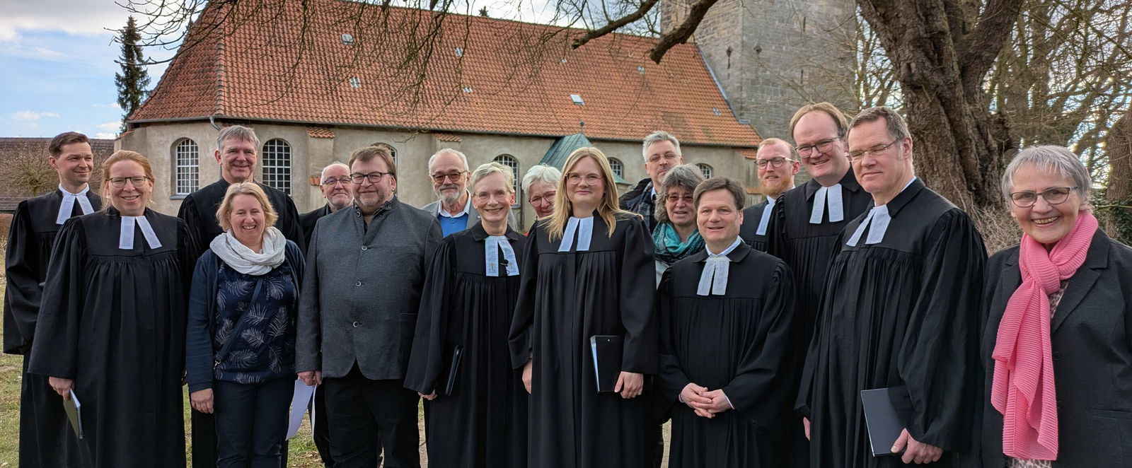 Gut ein Dutzend Personen, teils in Talaren, stehen unter einem Baum vor einer Kirche.