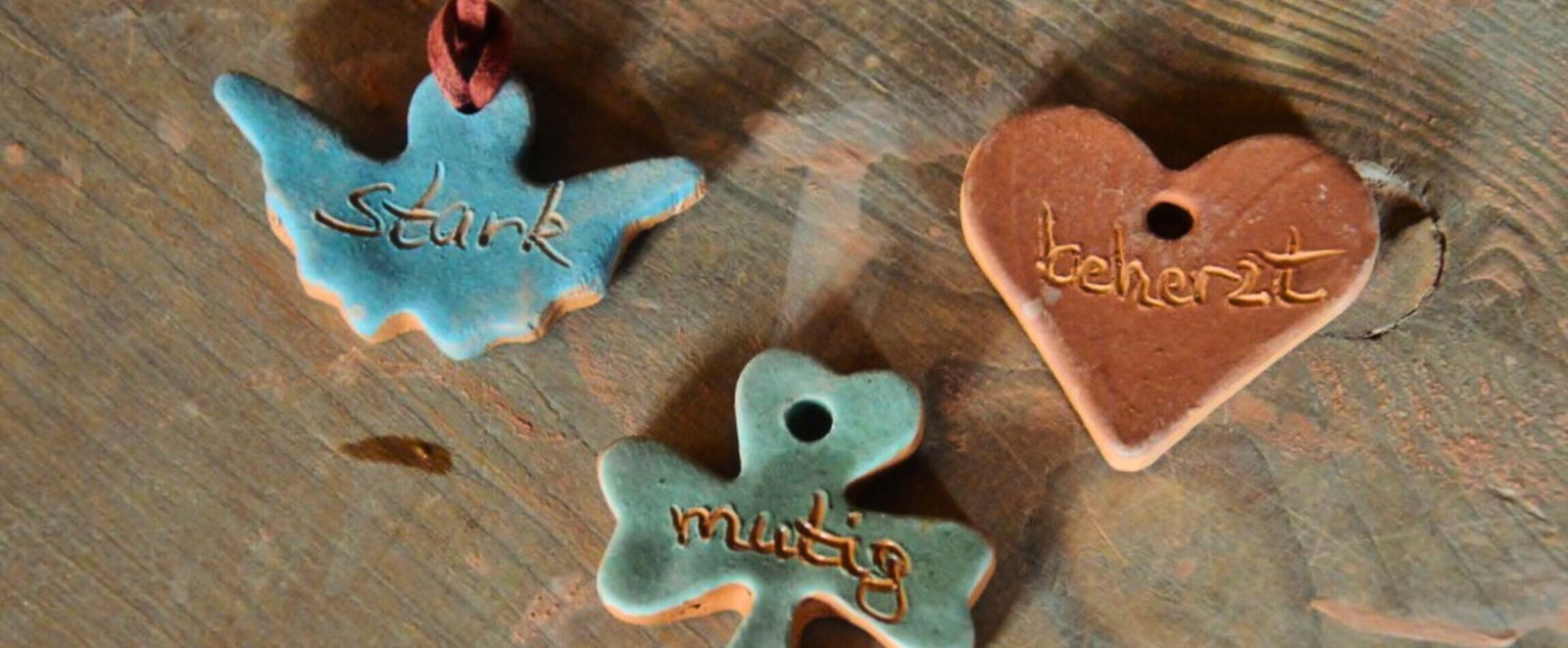 Drei Anhänger aus Ton, in Form eines Engels, Kleeblatts und Herzens. Darin steht mutig, stark und beherzt.