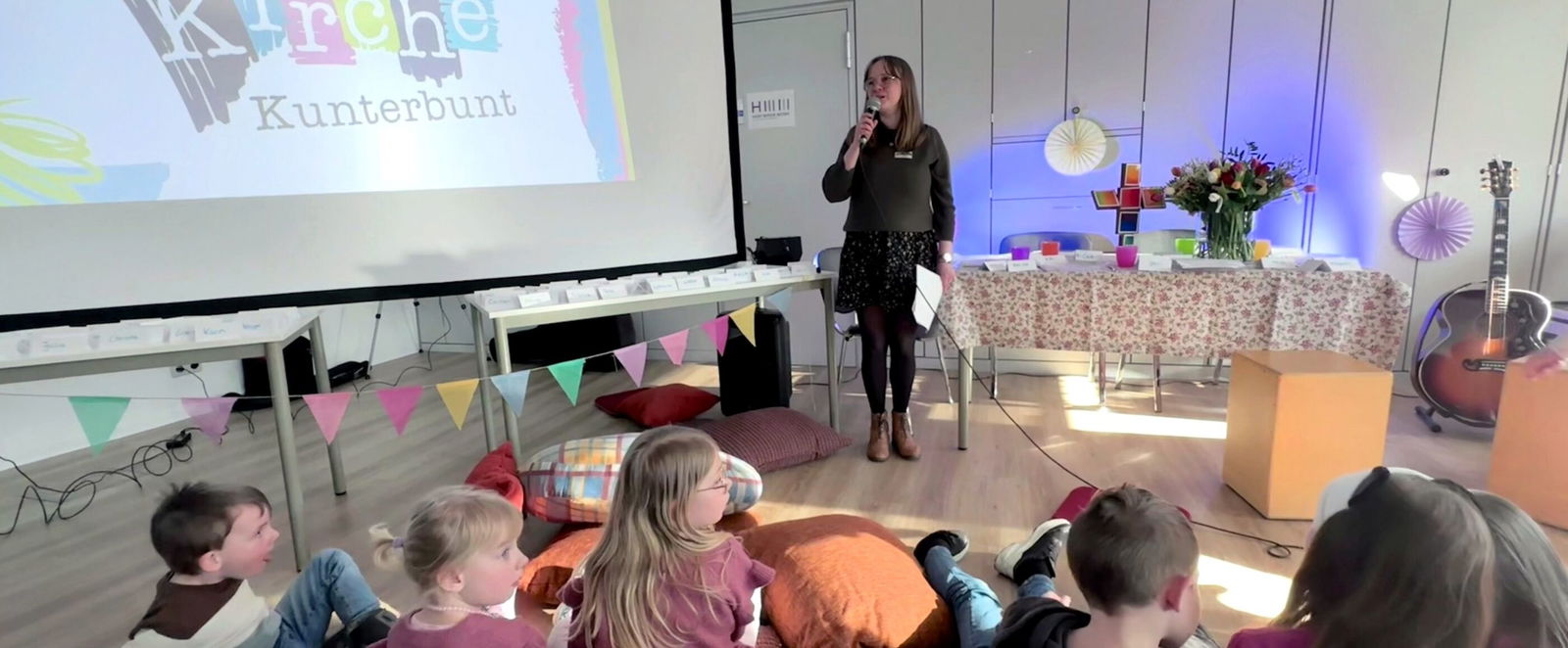 In einem Raum steht an einer Leinwand "Kirche kunterbunt", daneben eine weiblich gelesene Person mit einem Mikro. Vor ihr sitzen Kinder auf vielen bunten Kissen.