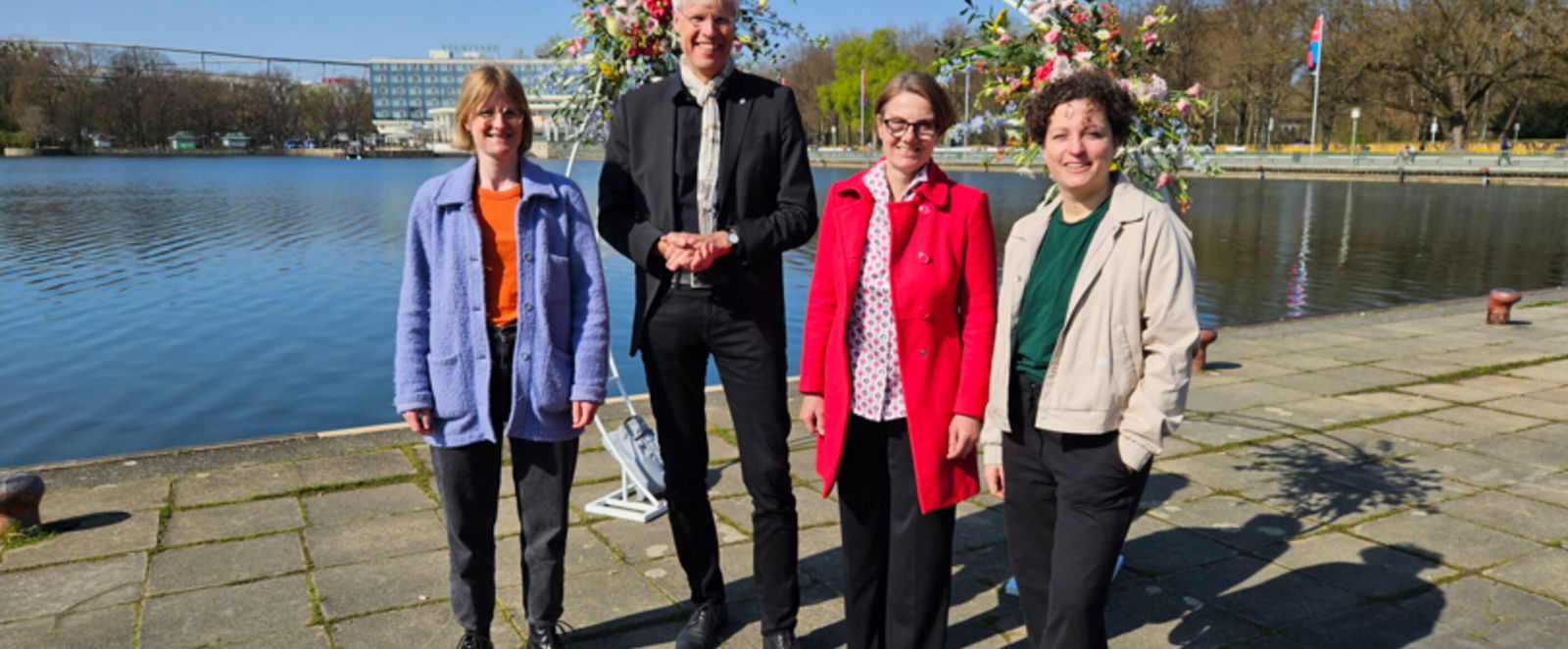 Eine Gruppe steht vor einem See. Drei davon sind als Frauen lesbar, einer als Mann.