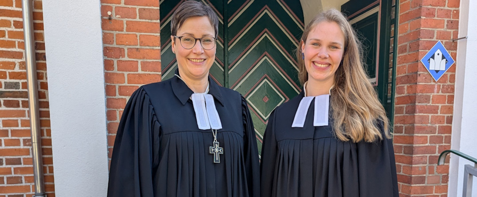 Zwei weiblich gelesene Personen in Talaren stehen vor einer Kirchentür.