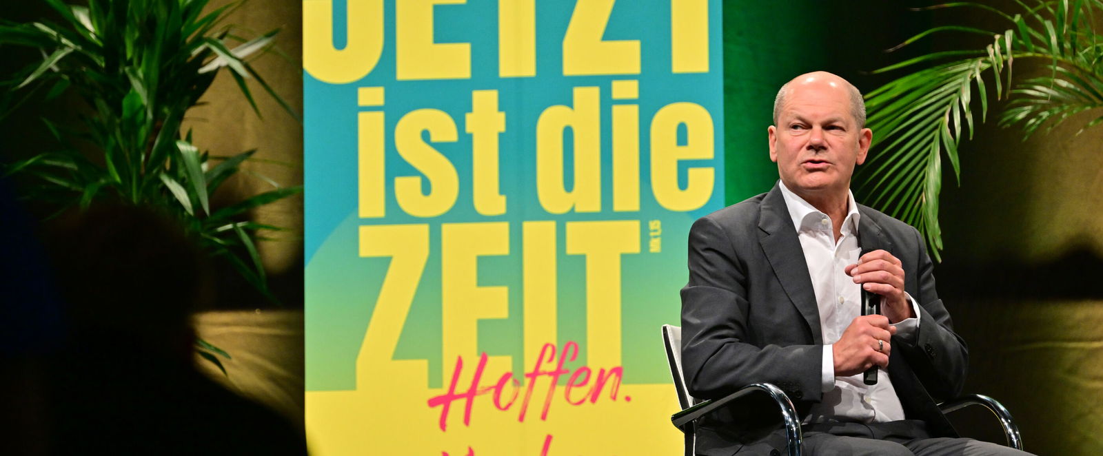 Ein Mann im Anzug sitzt auf einer Bühne, links neben ihm ein großes Roll-Up mit der Aufschrift „Jetzt ist die Zeit“