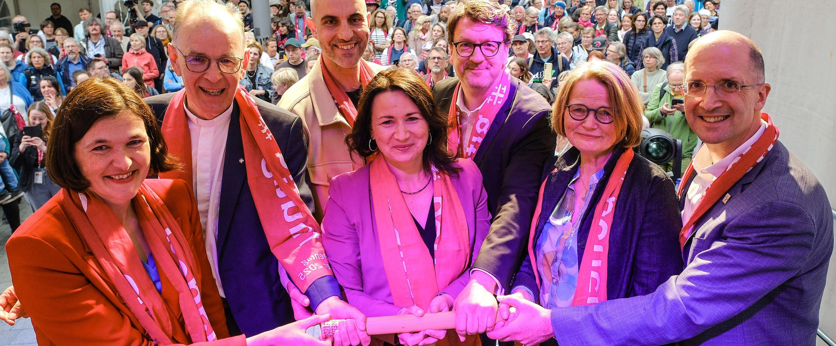 Mehrere Menschen halten einen Staffelstab in die Höhe. Manche tragen rote Schals.