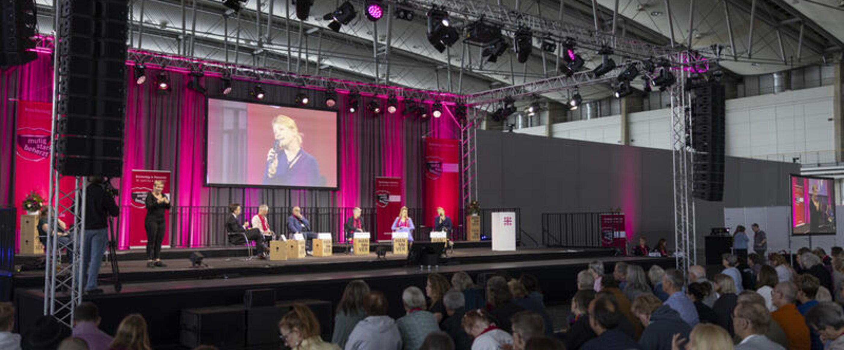 Auf einer rot erleuchteten Bühne diskutieren mehrere Personen miteinander, die im Halbkreis sitzen. Menschen sitzen in der Halle vor der Bühne und schauen zu.