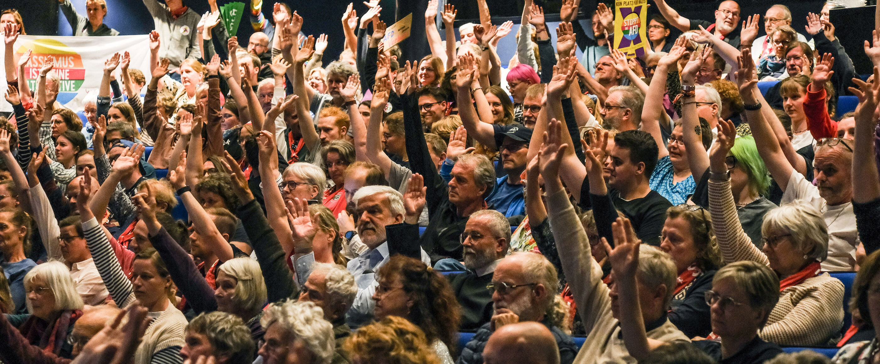 Viele Menschen sitzen in einem Publikumssaal und heben jeweils eine Hand.