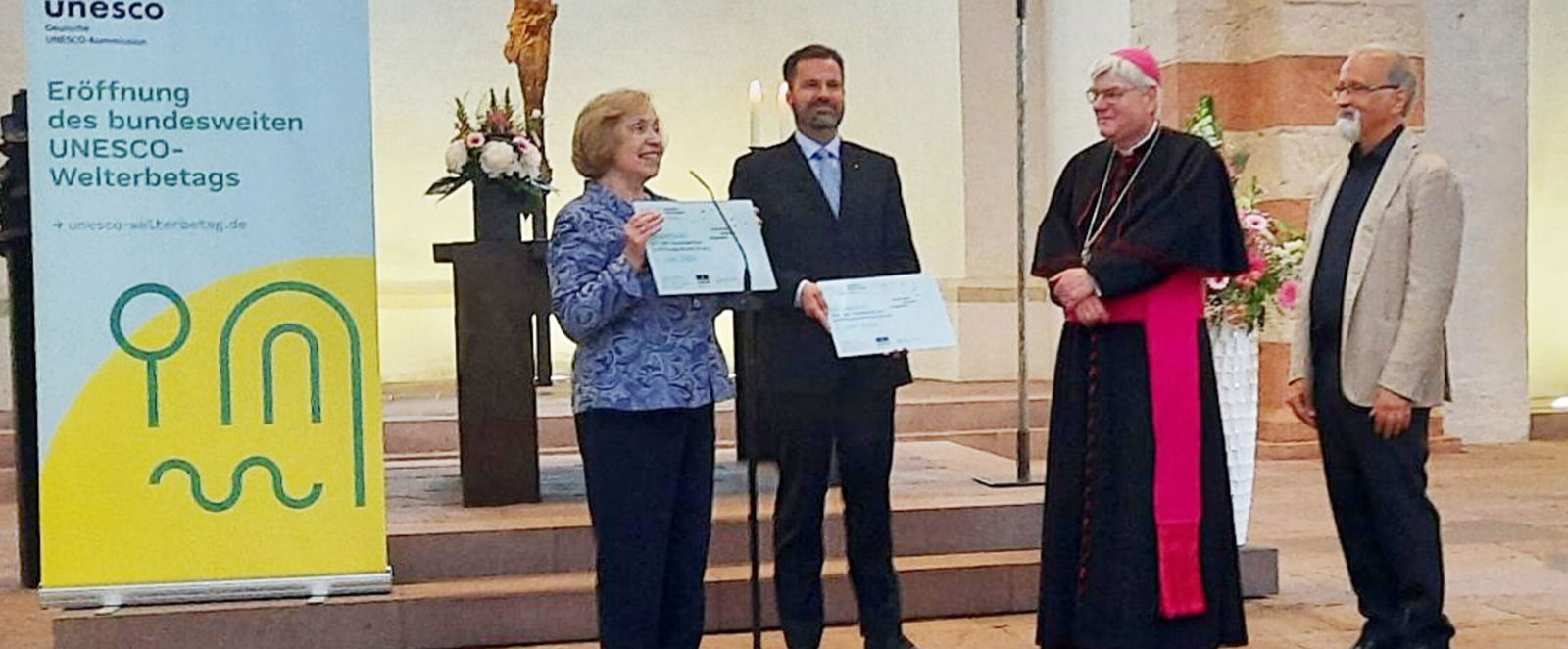 Eine als Frau lesbare Person und drei als Männer lesbare Personen stehen im Altarraum einer Kirche, zwei halten Schilder. Links steht ein Aufsteller mit der Aufschrift: Eröffnung des bundesweiten UNESCO-Welterbetags.