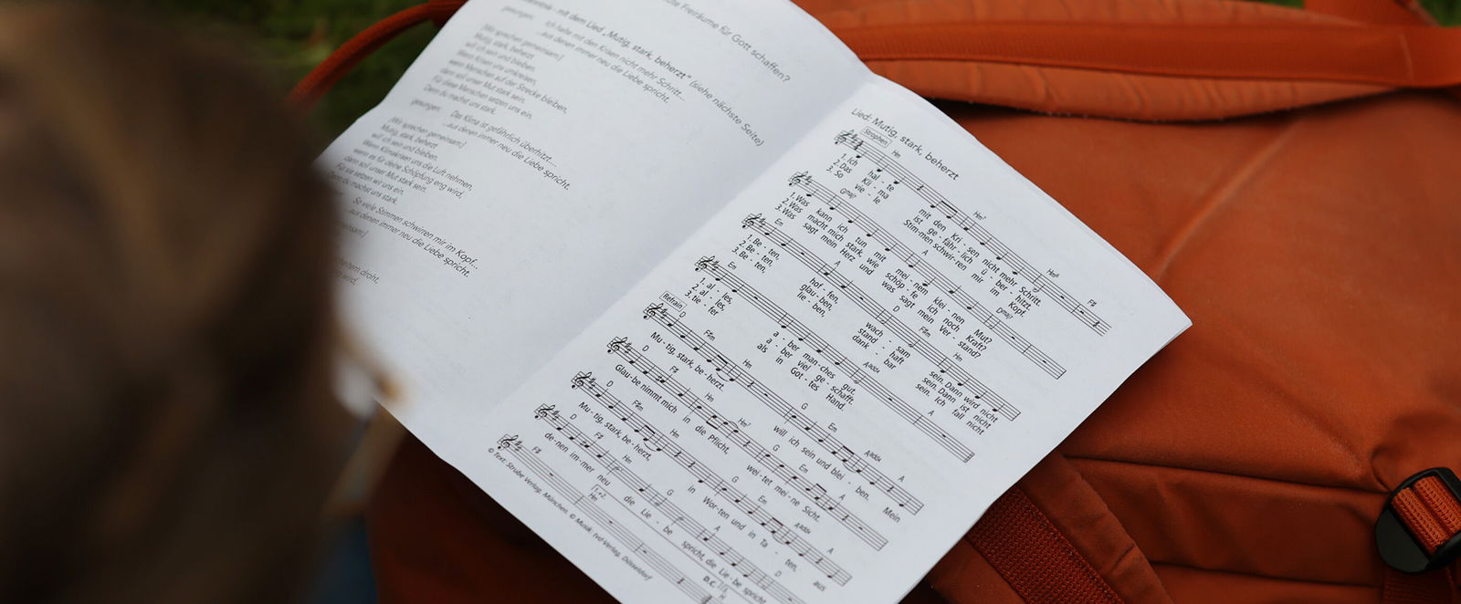 Eine Person singt aus einem Liedblatt, das auf einem Rucksack liegt.