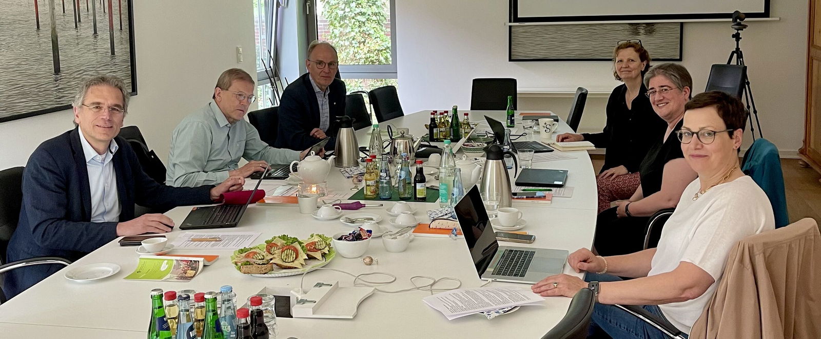 Eine Gruppe von sechs männlich bzw. weiblich gelesenen Personen sitzt an einem großen Konferenztisch; in der Mitte des Tisches liegen Arbeitsmaterialien sowie belegte Brötchen neben Getränken.