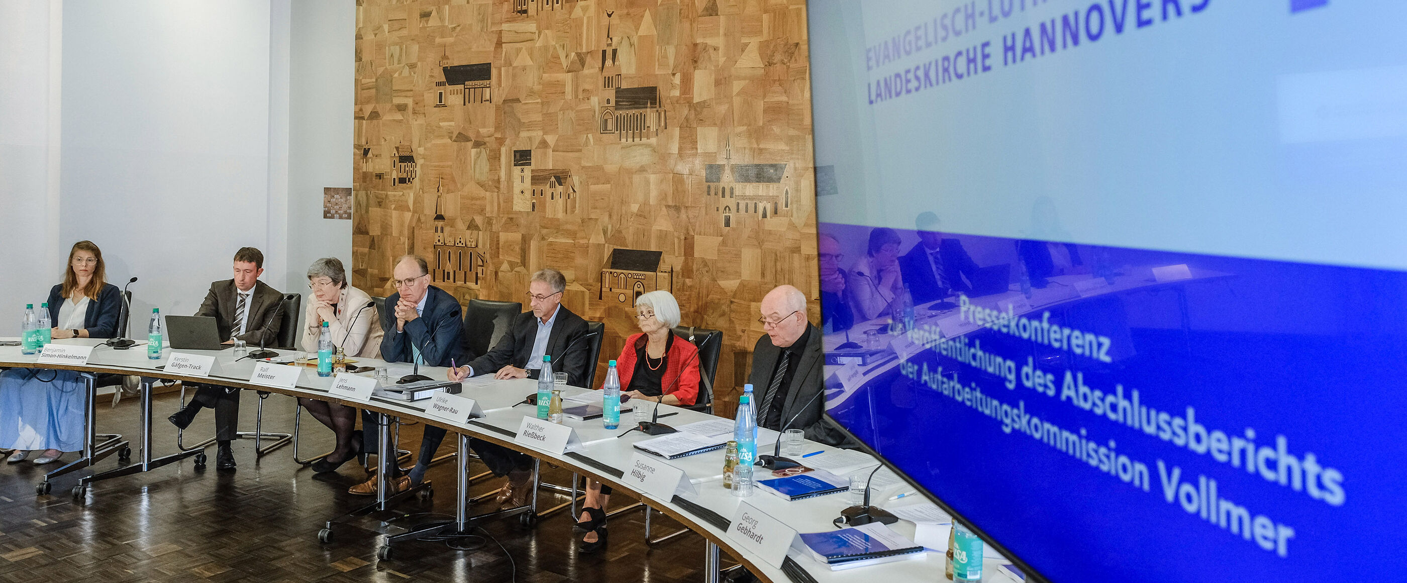 Im Vordergrund ein großer Bildschirm, im Hintergrund ein Podium mit als Frauen und Männer lesbaren Personen.
