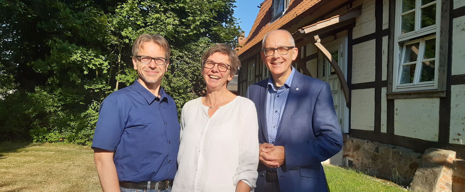 Zwei männlich und eine weiblich gelesene Person stehen vor einem Fachwerkhaus zusammen und lächeln in die Kamera.