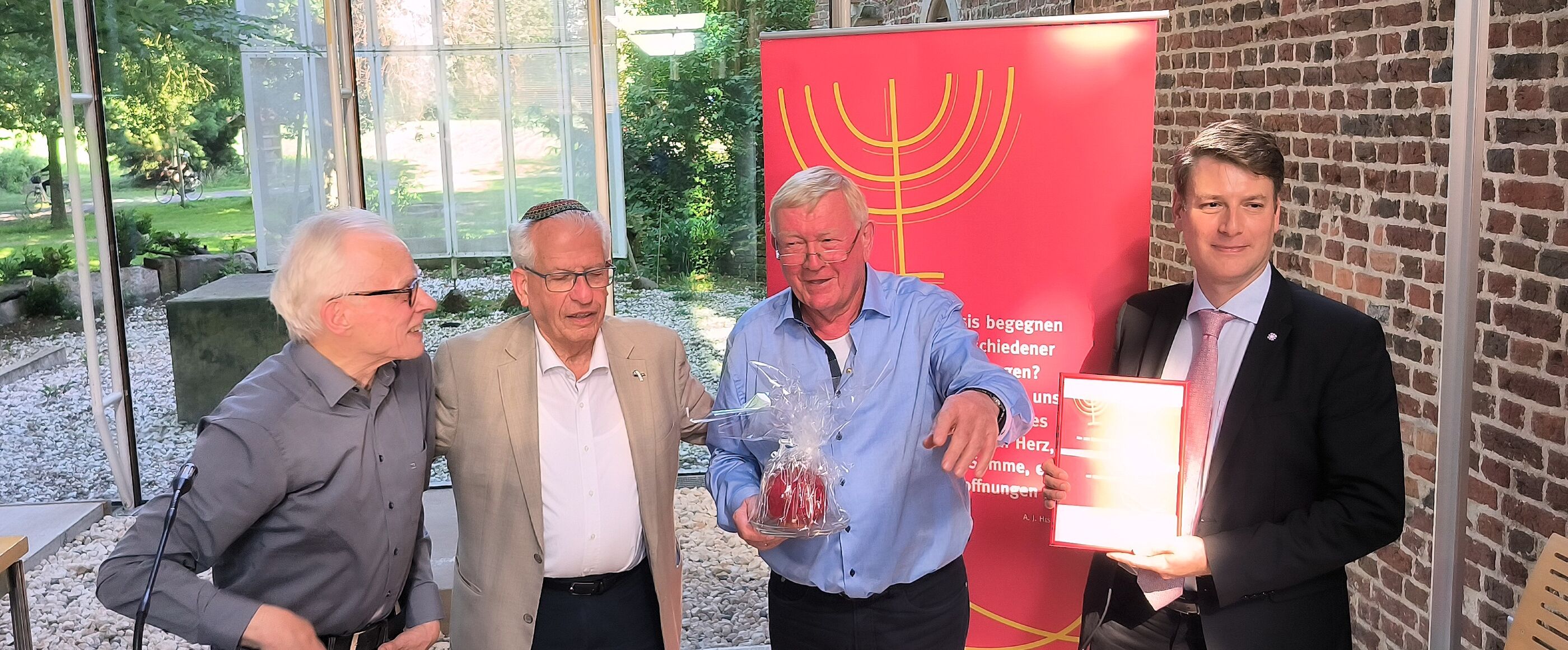 Vier männlich gelesene Menschen stehen zusammen, einer trägt eine Kippa. Einer hält eine Auszeichnung in Händen, ein anderer ein Geschenk.