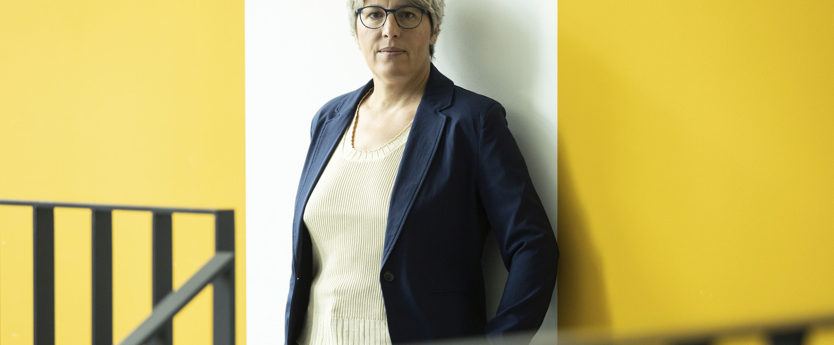 Eine weiblich gelesene Person mit kurzen, blonden Haaren vor einer weiß-gelben Wand.
