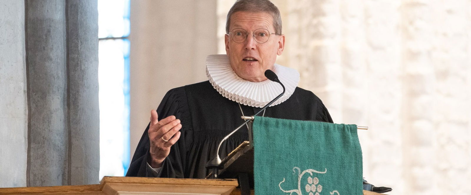 Eine männlich gelesene Person mit kurzen Haaren, in Talar, steht auf einer Kanzel an einem Mikro.
