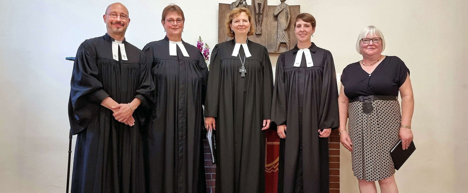Vier Personen in Talaren und eine Person in Bluse und Rock stehen nebeneinander vor einem Altar.