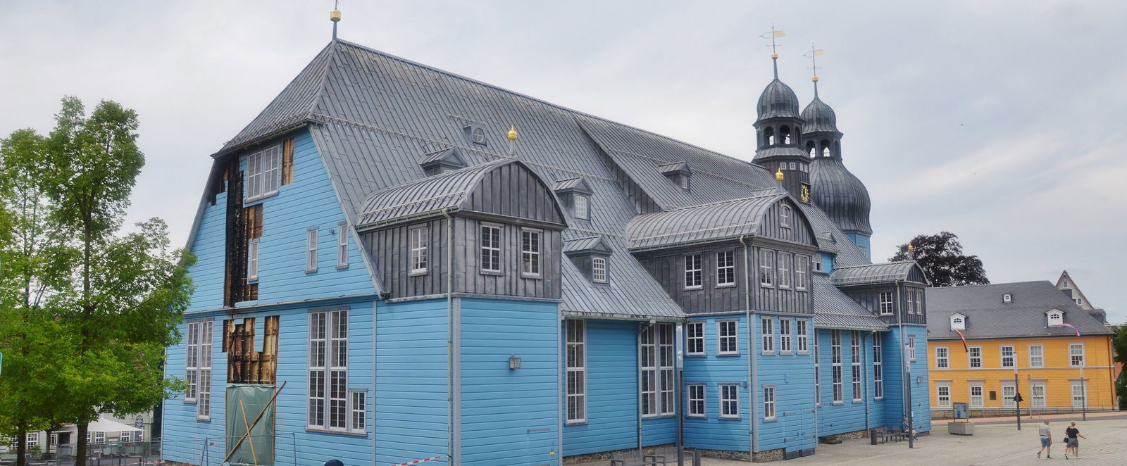 Ein hellblaues Holzgebäude (eine Kirche), deren Fassade an der kurzen Seite beschädigt ist. 