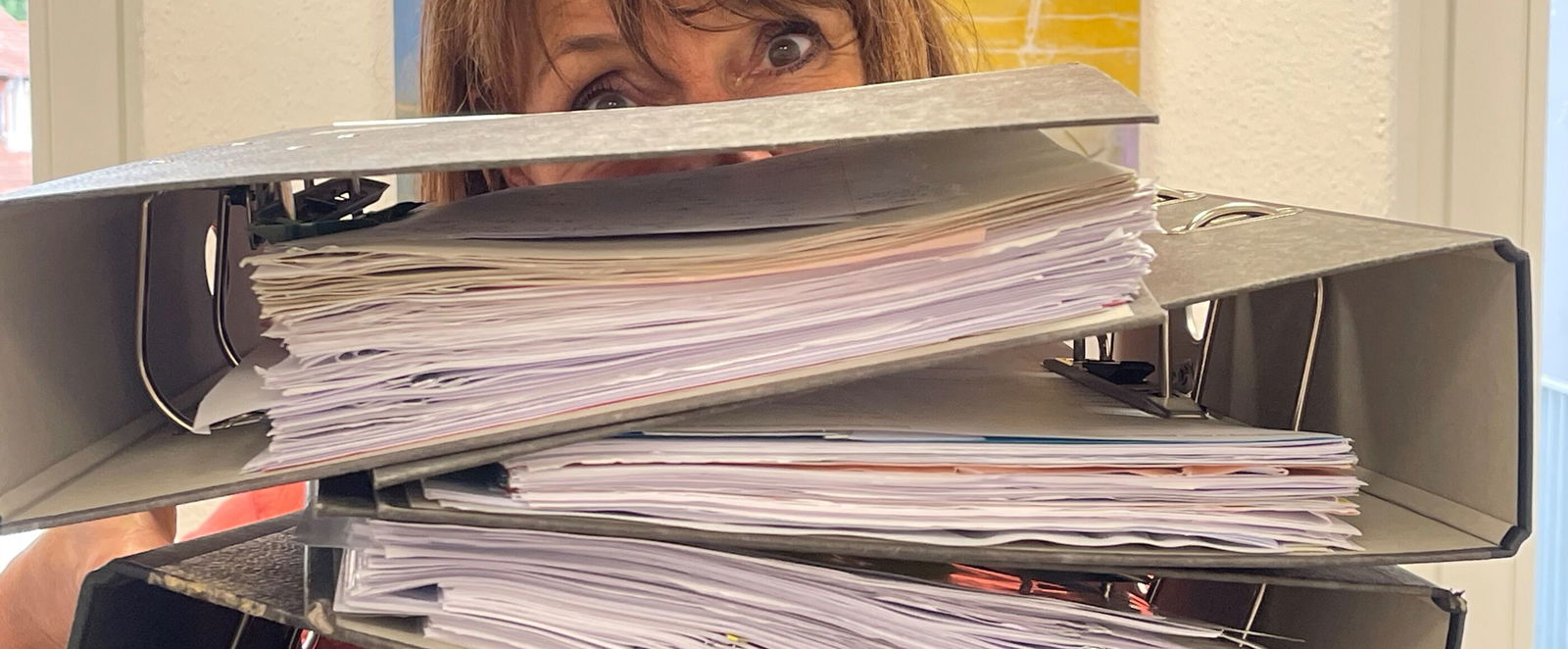 Eine weiblich gelesene Person, die hinter dicken Akten hervorschaut.