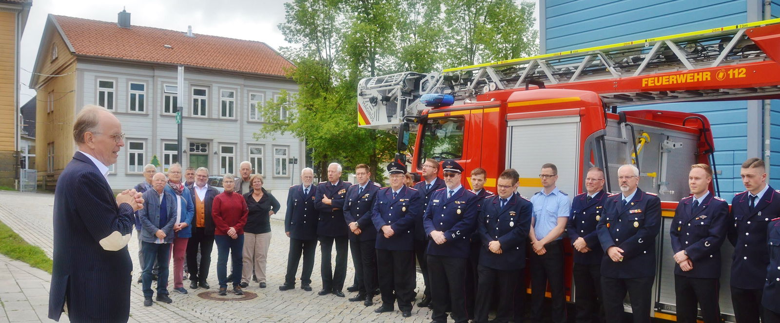 Mehrere Feuerwehrleute stehen vor einem Feuerwehrwagen und schauen eine männlich gelesene Person in heller Hose und dunklem Jackett an.