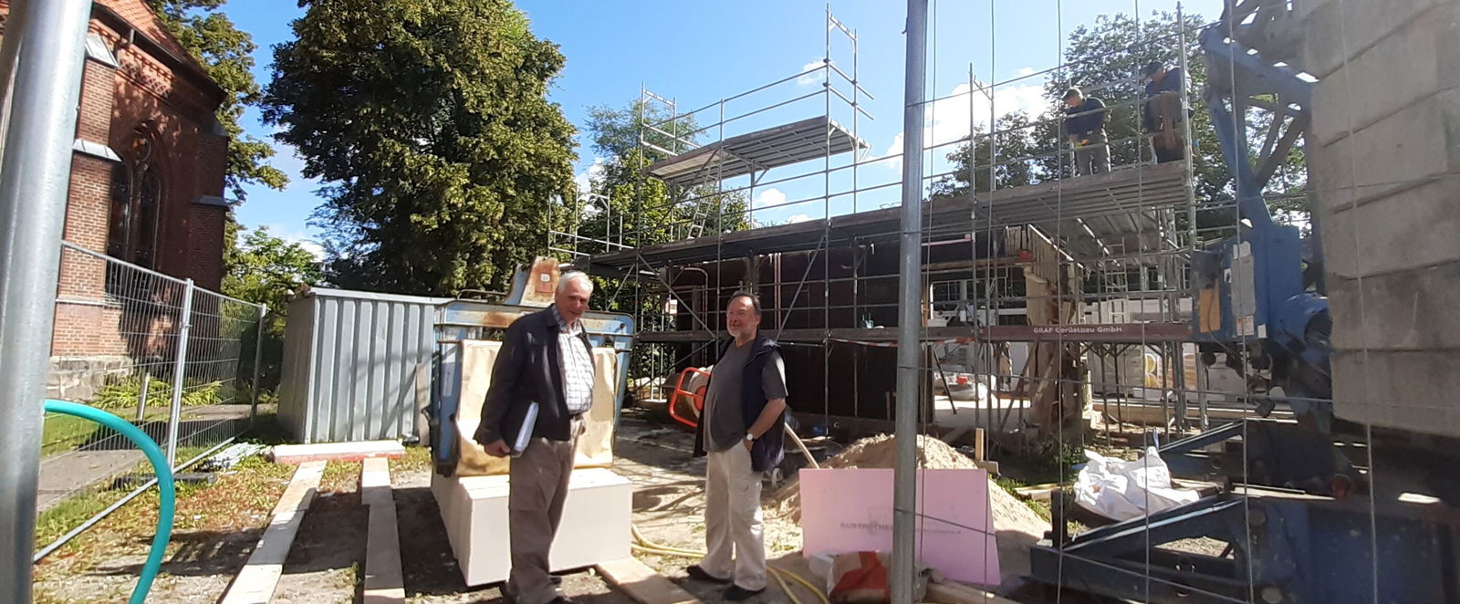 Mehrere Menschen stehen auf einer Baustelle, ein größeres Haus ist im Rohbau zwischen Bauzaun und Materialien zu erkennen.