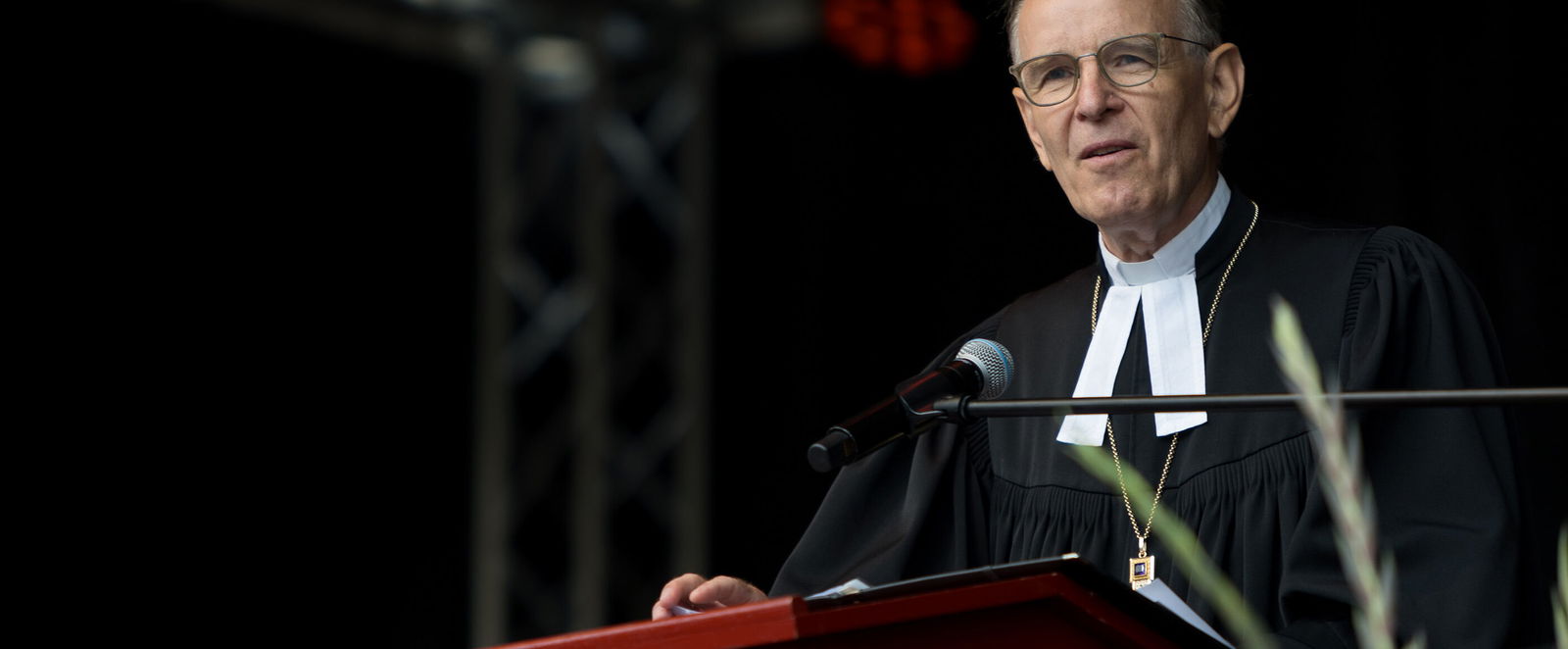 Eine mittelalte als Mann lesbare Person steht im Talar an einem Redepult und spricht.