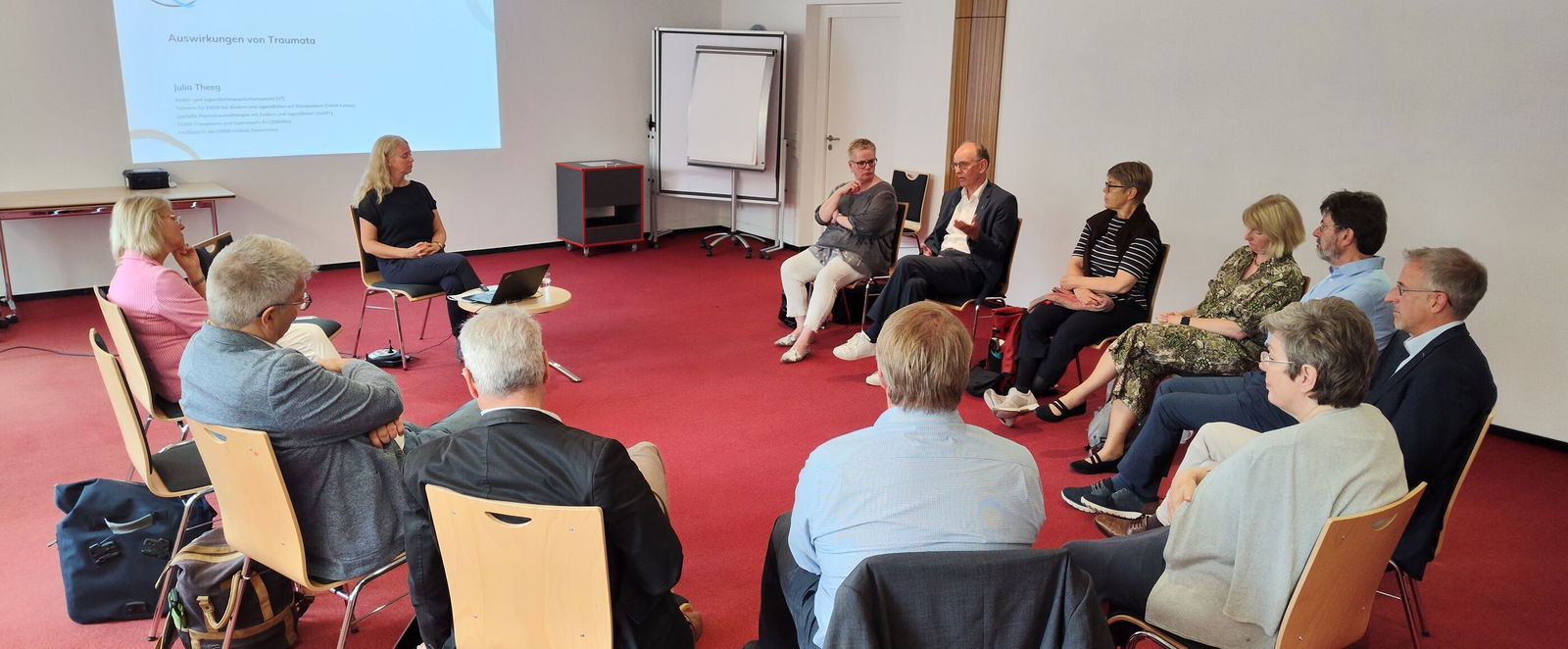 In einem Konferenzraum sitzen mehr als ein Dutzend Personen auf Stühlen in einem Kreis im Gespräch.