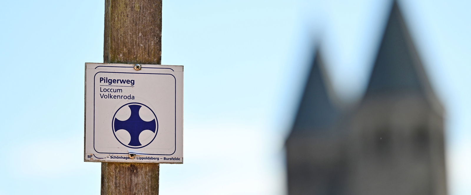 Ein Schild mit der Aufschrift Pilgerweg Loccum-Volkenroda; im Hintergrund ist eine Kirche mit zwei Türmen zu sehen.