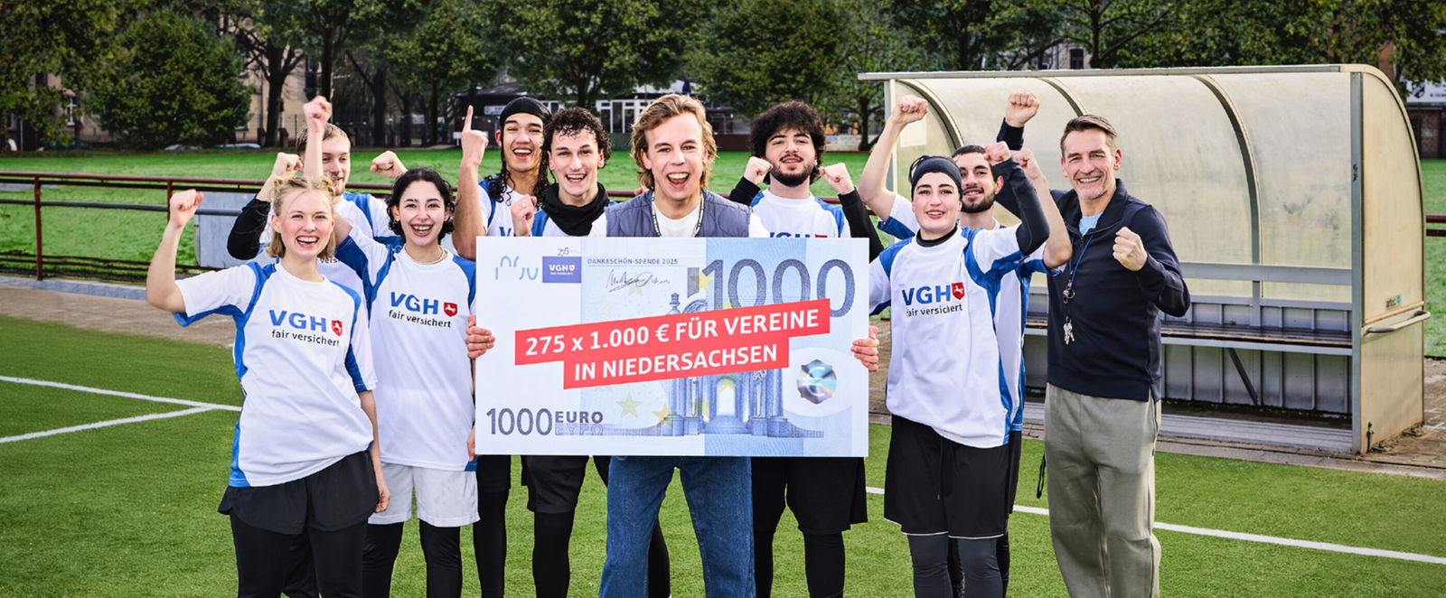 Eine Gruppe aus jungen Menschen steht auf einem Sportplatz und hält jubelnd einen symbolischen Scheck hoch.