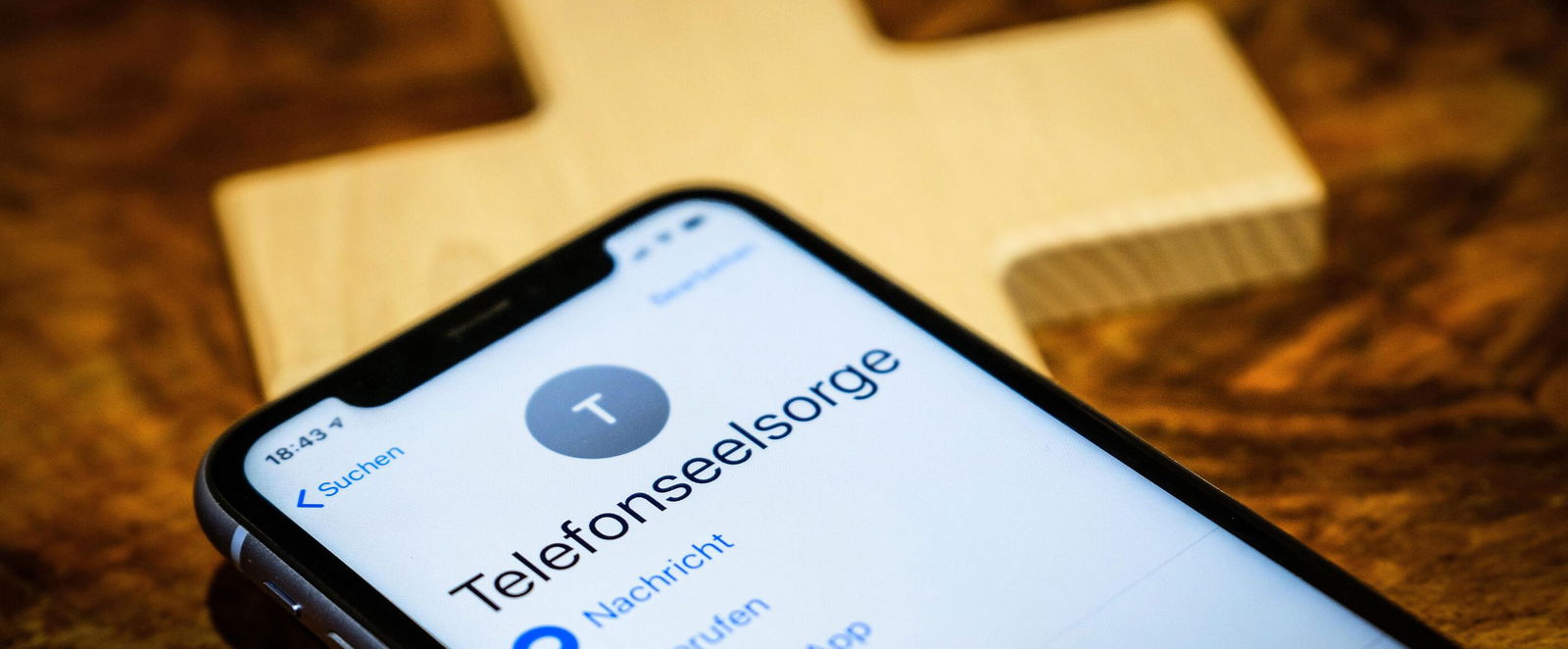 Ein Mobiltelefon, auf dessen Display "Telefonseelsorge" steht. Darunter liegt ein Holzkreuz.