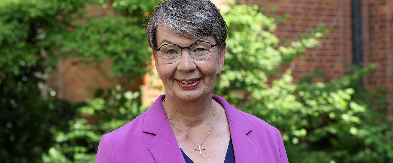 Eine als Frau lesbare Person mit violettem Blazer steht in einem Garten.