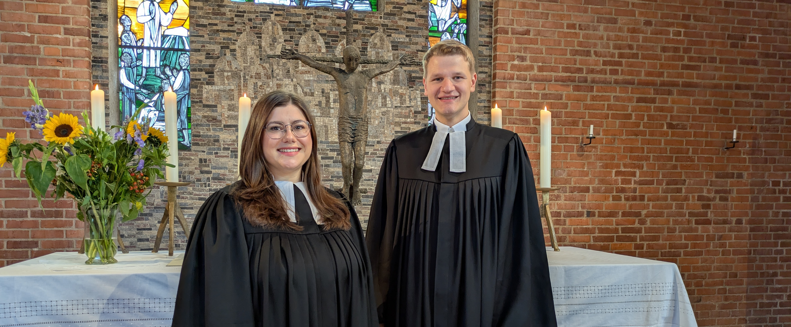 Eine weiblich und eine männlich gelesene Person in Talaren stehen vor einem Altar in einer Kirche.
