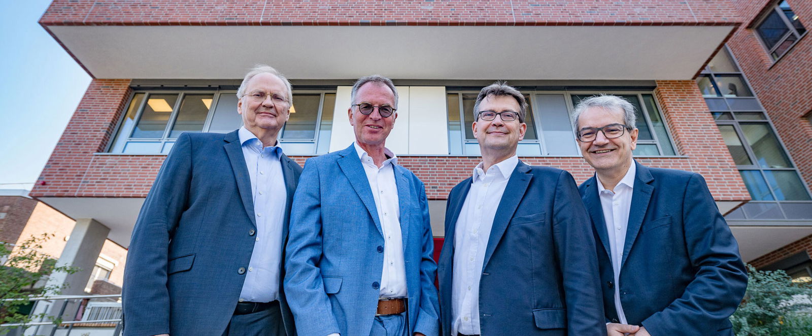 Vier als Männer lesbare Personen im Anzug posieren vor einem Gebäude.