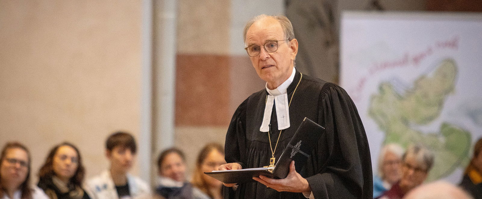 Eine als Mann lesbare Person im Talar spricht in einer Kirche vor Menschen.