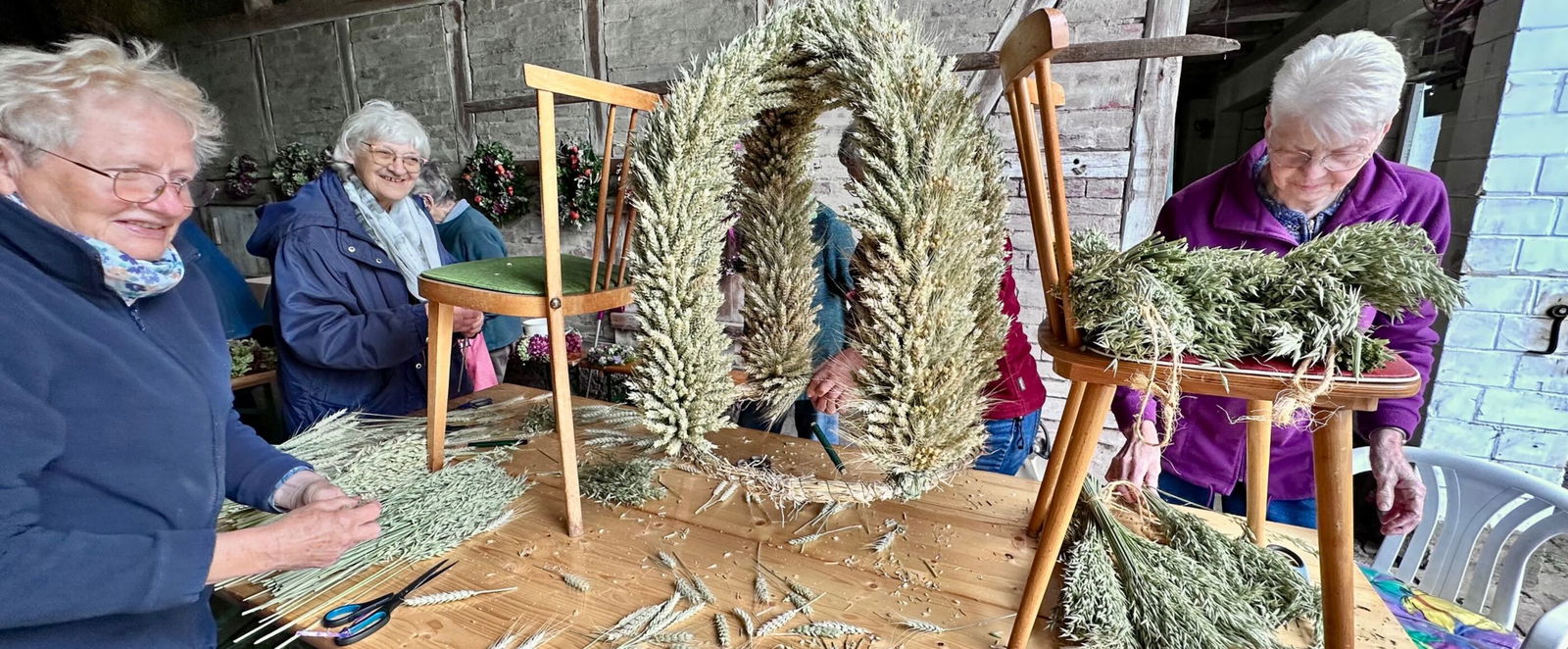 Drei Personen stehen an einem Tisch, eine Erntekrone entsteht in der Mitte.