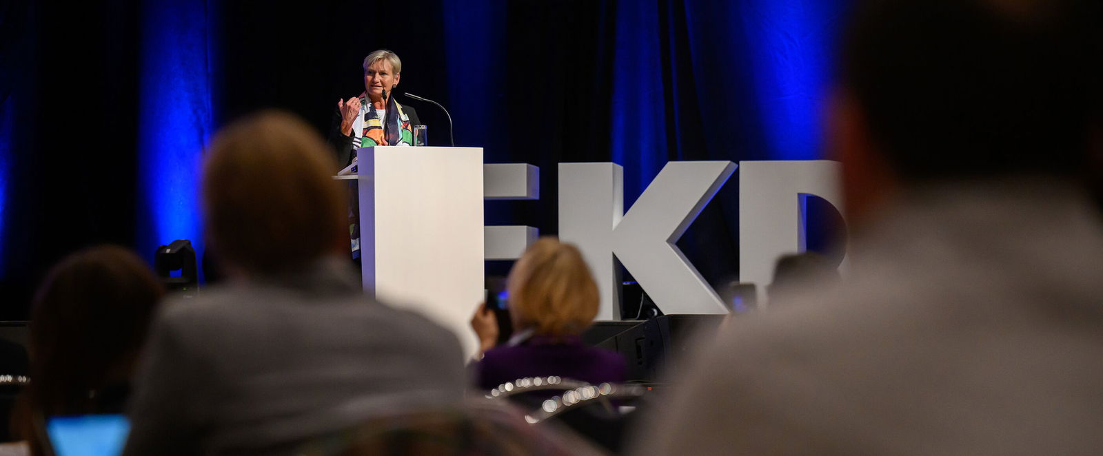 Eine als Frau lesbare Person steht auf einer Bühne am Redepult.