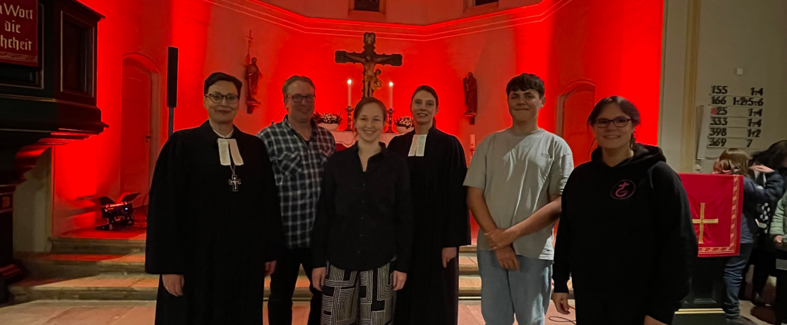 Sechs Personen, zwei im Talar, stehen in einer rot beleuchteten Kirche.