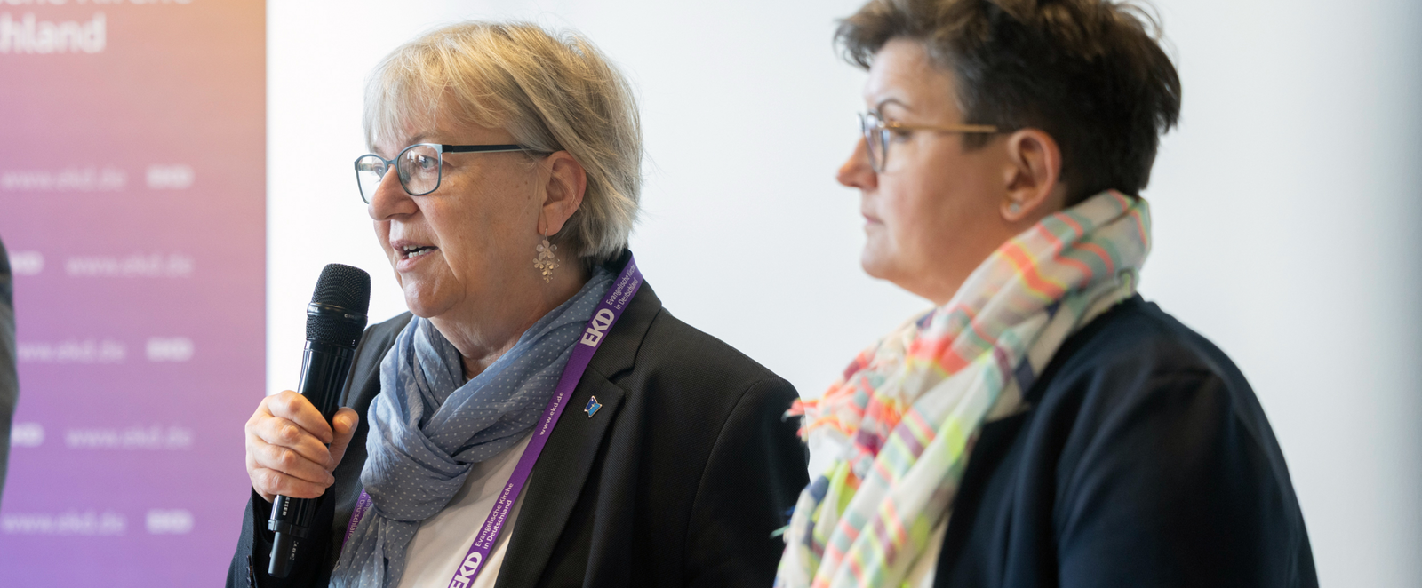 Zwei weiblich gelesene Personen mit einem Mikro.