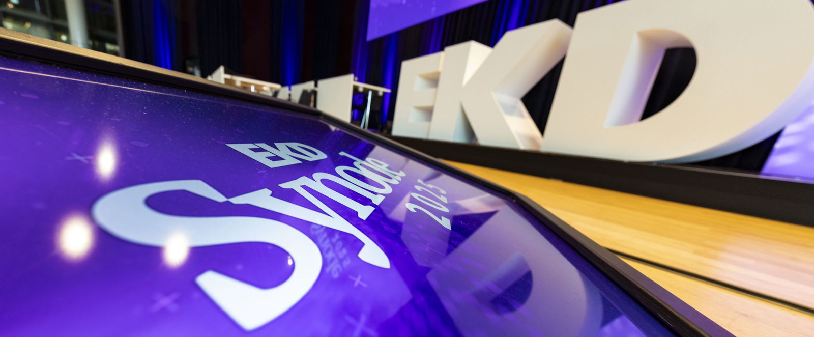 Ein Bildschirm zeigt: Synode. Im Hintergrund drei übergroße Buchstaben auf einer Bühne: EKD