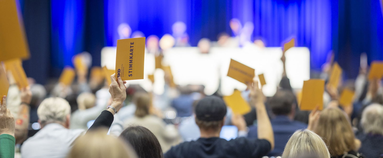 Menschen in einem blau beleuchteten Saal heben gelbe Karten zur Abstimmung.