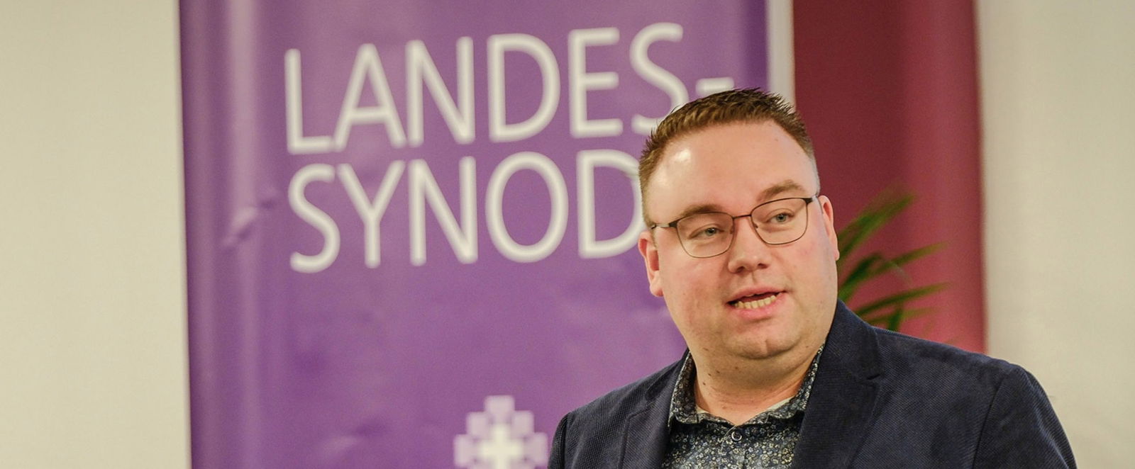 Ruben Grüssing vor dem lila Banner der Landessynode.