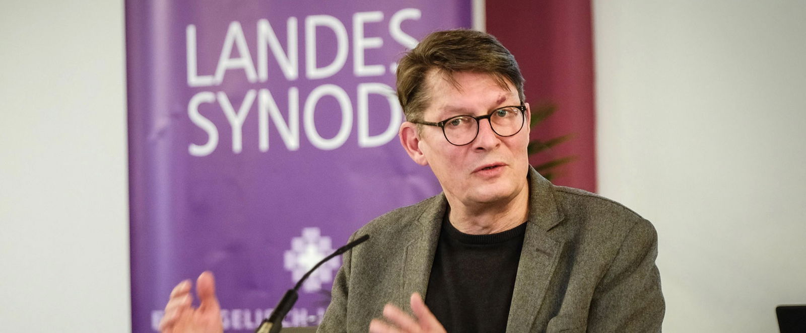 Martin Krarup steht am Rednerpult vor dem lila Banner mit der weißen Schrift "Landessynode".